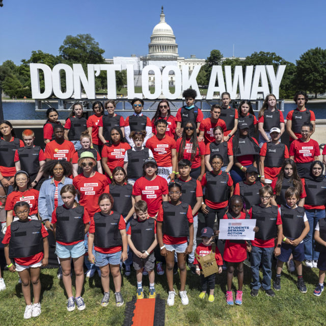 A group of students wearing bullet proof vests and Students Demand Action shirts stand in front of a sign that says Don't Look Away with the Capitol building in the background during an event in Washington, DC in June 2022