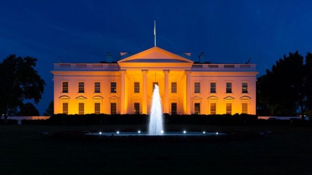 A front-facing view of the White House taken at night. A fountain is bubbling in the center of the shot, bisecting the White House. The White House is lit up the color orange for gun violence awareness. The sky behind it is navy blue.
