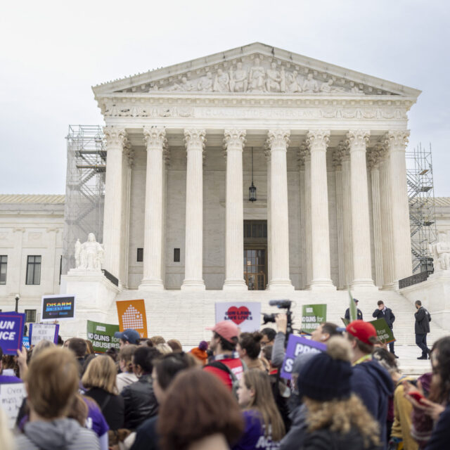 Gun violence prevention advocates rally outside of the Supreme Court. The bottom third of the image shows people in winter clothing—hats, sweatshirts—holding signs that read 