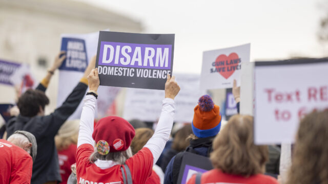 A view from the back of a crowd of Moms Demand Action volunteers protesting in front of the Supreme Court. One volunteer holds a sign that reads 