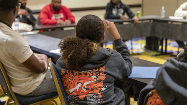 A group of people sit around a rectangular table at the 2023 Young Black Changemakers Summit. One participant is in focus; she is photographed from behind, and is wearing a sweatshirt that reads 