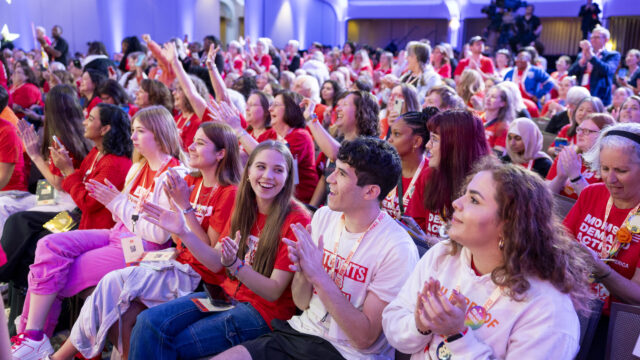 Students Demand Action volunteers smile and clap in the front row of an auditorium. They are wearing lanyards that read 