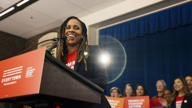 Angela Ferrell-Zabala, the executive director of Moms Demand Action and Students Demand Action, smiles while speaking at a podium. She wear metal hoop earrings, a black blazer, and a red Moms Demand Action t-shirt. Behind her, a cluster of 7 volunteers hold Moms Demand Action and Everytown placards and clap as she talks.