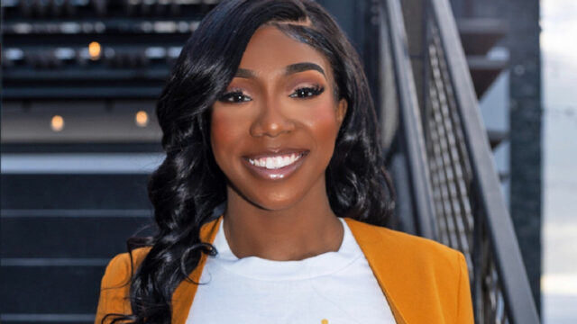 Aaliyah Strong, the founder and executive director of Tyme to Thrive Beyond Grief, poses for a portrait photo. Her black hair is parted on the side and she is wearing subtle glamorous makeup. She is facing the camera directly and is pulling her orange blazer slightly away from her white t-shirt so the full 