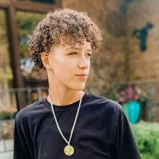Alex Franco, a trans man, stands with his body facing the camera and his face turned at a profile angle. He wears a black long-sleeved shirt and a metal necklace with what appears to be a gold coin hanging from it. His hair is cropped to around his ears and is curly.