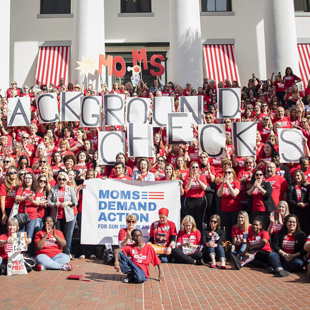 A large group of Moms Demand Action volunteers hold up signs spelling, 'Background Checks' at an Advocacy Day in Florida