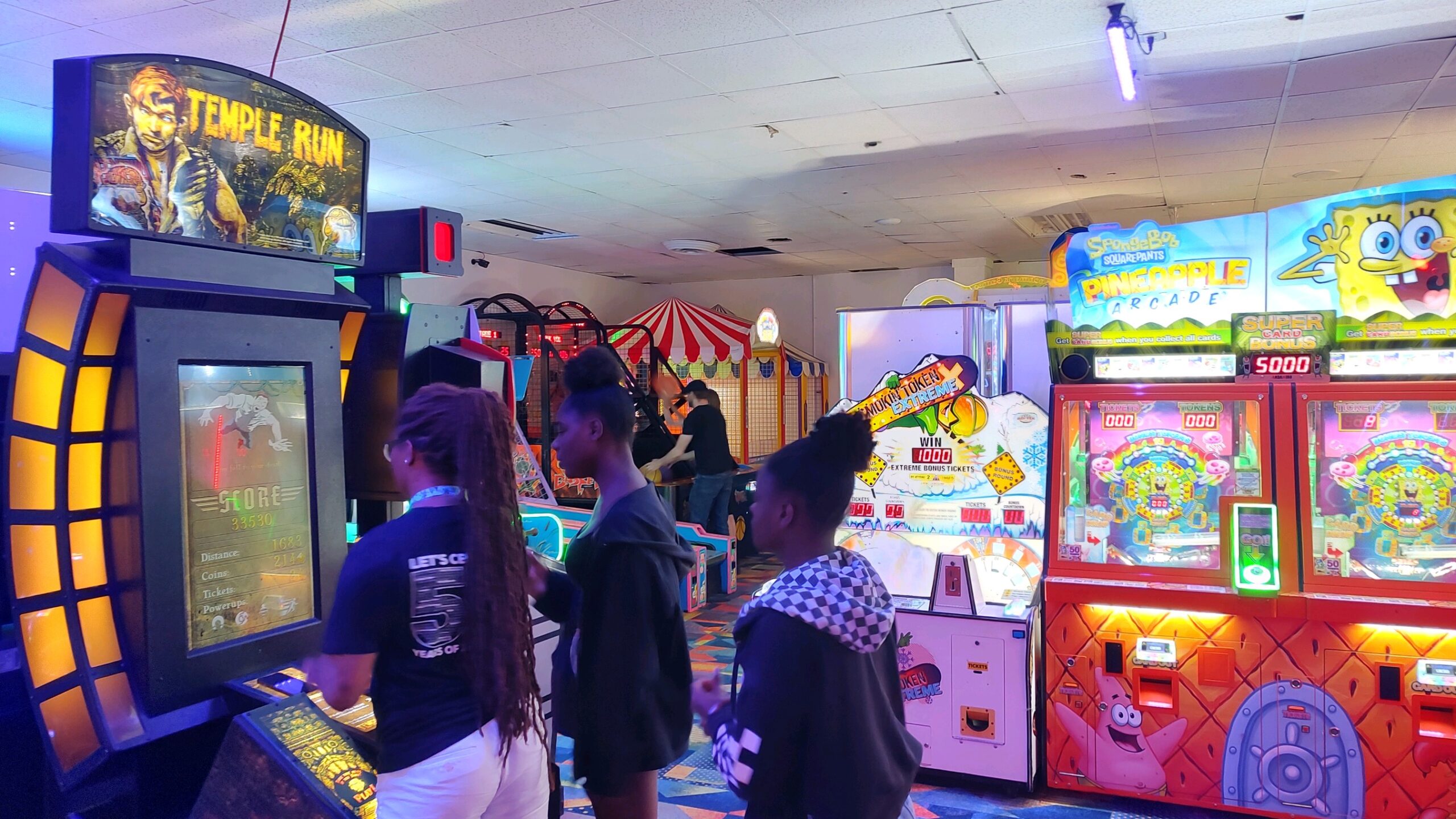 Three people are seen at a side profile. They are standing in front of arcade games at a bowling alley.