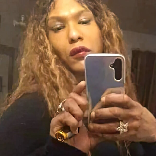 Diamond Brigman, a Black trans woman, holds her phone with both hands as she takes a photo in a mirror. She is wearing chunky gold rings on each hand; her hair is long, curly, and parted down the middle.