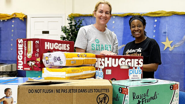 Shannon, a white woman with blond hair wearing a grey Be SMART t-shirt, poses with Crystal, a Black woman wearing a black t-shirt and a black headband. In the foreground of the image are boxes of diapers they have collected.