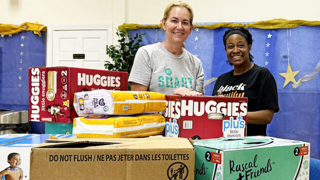 Shannon, a white woman with blond hair wearing a grey Be SMART t-shirt, poses with Crystal, a Black woman wearing a black t-shirt and a black headband. In the foreground of the image are boxes of diapers they have collected.