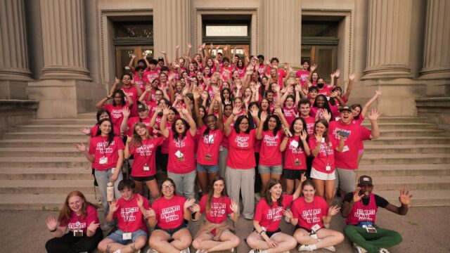 A group of 100 Students Demand Action volunteers, all wearing red t-shirts with the Students Demand Action logo, gather for a photo on the Vincent Hall steps at the University of Minnesota during the Summer Organizing Institute. Students are raising their arms in the air and waving at the camera.
