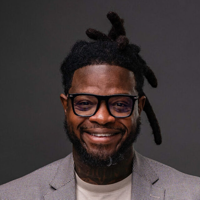 A portrait photo of DuJuan Kennedy, who is posing in front of a professional charcoal-gray background. He wears glasses with a thick black frame, a cream-colored t-shirt, and a thin plaid-stripe blazer.