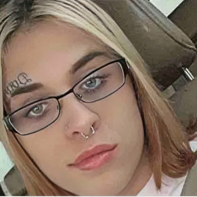 Dylan Gurley, a trans woman, takes a selfie while sitting in the car. She wears black rectangular glasses and has a metal loop septum piercing; she has a script-font tattoo above her right eyebrow. Her blonde hair is shoulder-length and parted down the middle; she wears a white shirt and has a seatbelt over her left shoulder.