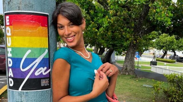 Joanna Cifredo, a human rights activist, smiles while leaning on a pole that has a poster with a rainbow pride flag on it