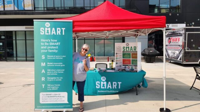 A Be SMART volunteer wears black pants and a grey t-shirt with the teal Be SMART logo. She stands next to a teal Be SMART standard. Behind her is a table covered with a teal tablecloth with the Be SMART logo and a red canopy to give shade while tabling.