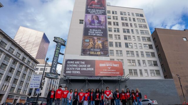 18 Students Demand Action volunteers pose for a low-angle photo. All are wearing red Students Demand Action t-shirts, pants, and jackets or long-sleeve layers. Behind the students, an 
