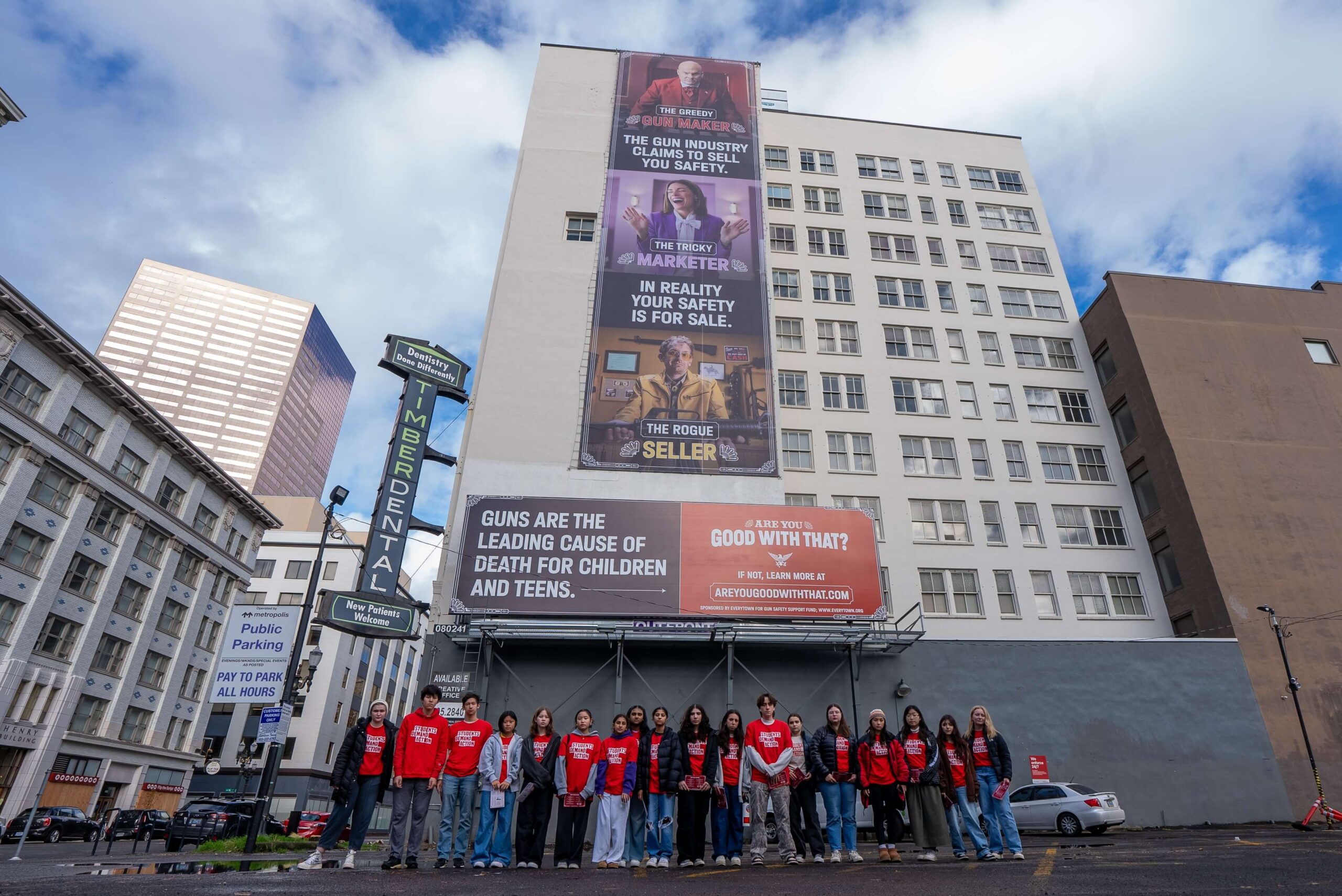 18 Students Demand Action volunteers pose for a low-angle photo. All are wearing red Students Demand Action t-shirts, pants, and jackets or long-sleeve layers. Behind the students, an "Are You Good With That" billboard is displayed on the side of a 10-story building. From top to bottom, it reads "The gun industry claims to sell you safety. In reality your safety is for sale. Guns are the leading cause of death for children and teens. Are you good with that?"