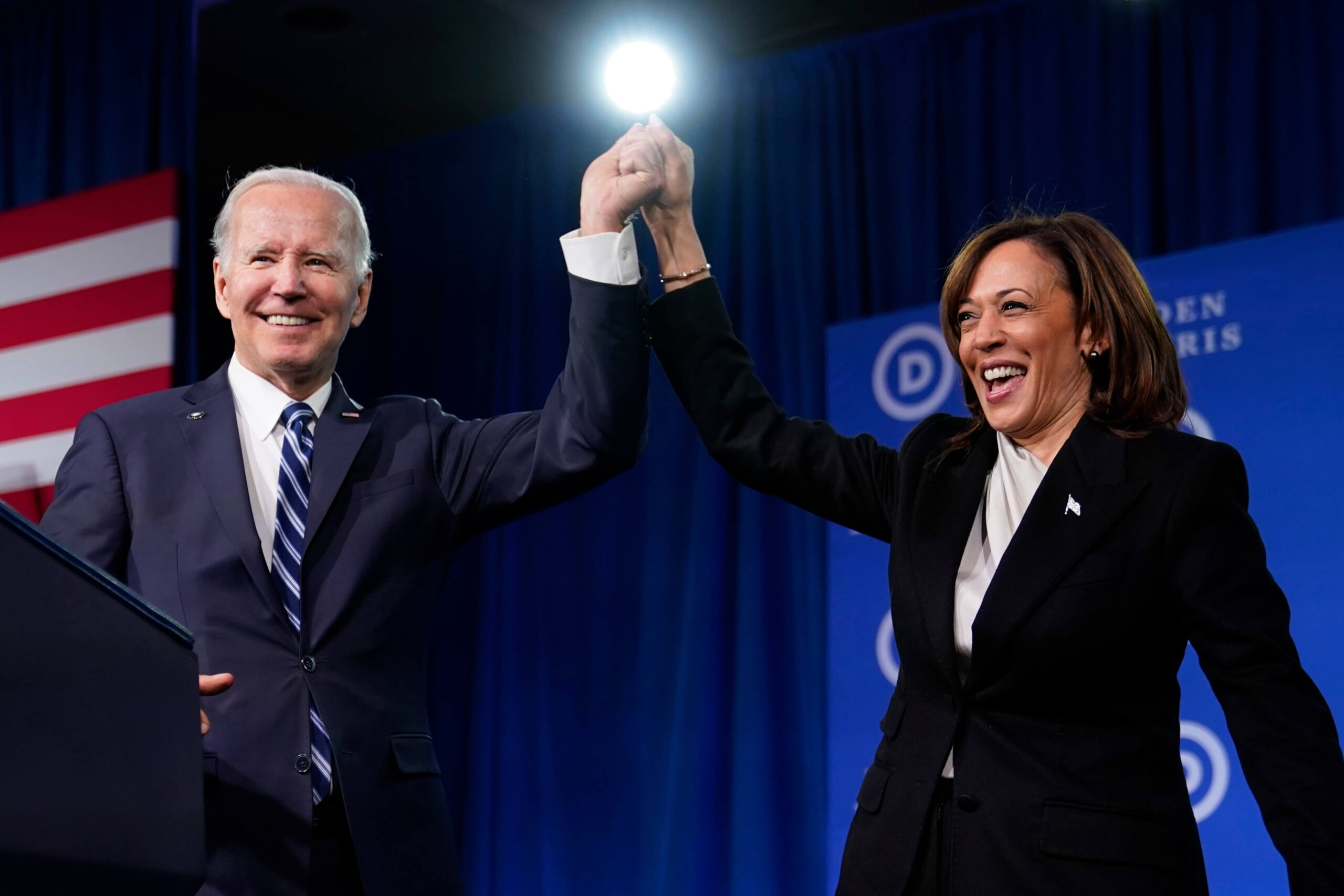 Joe Biden and Kamala Harris raise their joined hands as they stand behind a podium and smile at the crowd.
