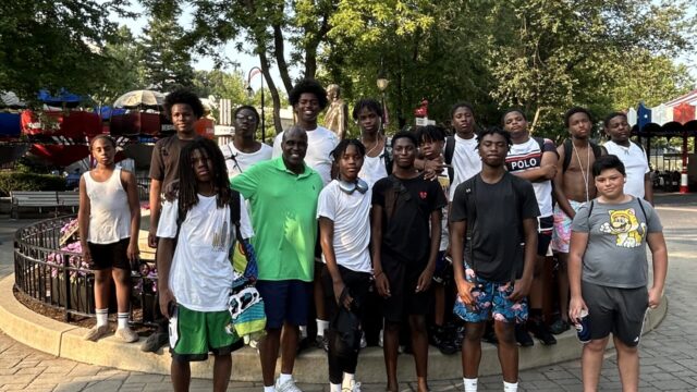 A group of 15 mentees post with a ManUpPHL mentor. They appear to be at an amusement park. The mentees are boys between the ages of 13 and 17.
