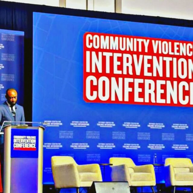 Michael-Sean Spence, a Black man wearing a blue suit, light collared shirt, and a tie, presents at a podium in front of a screen that reads 