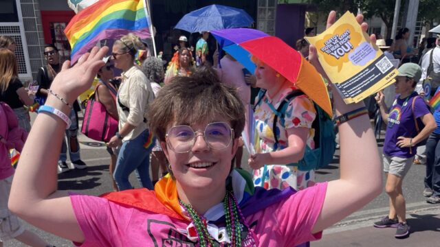 Bo Wilson wears a pink t-shirt, a red cape, and is holding pride flags at a Pride event.