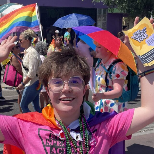 Bo Wilson wears a pink t-shirt, a red cape, and is holding pride flags at a Pride event.