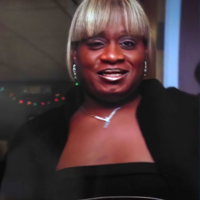 Kita Bee, a Black trans woman, takes a selfie; her blond hair is cut in a chin-length bob with bangs that hang straight across her forehead. She has bright silver earrings and a light necklace on; she is wearing a dark top.