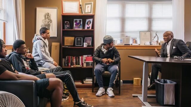 Leonard Jahad, the founder and executive director of Connecticut Violence Intervention and Prevention, is sitting at a desk to the far right of the image. He is wearing a grey suit and a white collared shirt. He is speaking with 4 CTVIP participants, 3 of whom sit on a couch and one of whom sits on a chair around his office.
