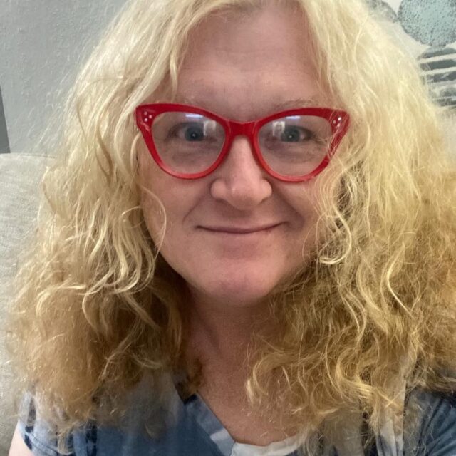 Meghan Riley Lewis, a white trans woman, smiles for a selfie. She has shoulder-length curly blond hair and wears red cat-eye glasses.