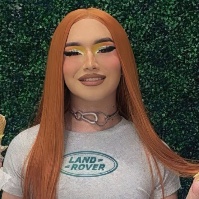 Meraxes Medina, a Latina trans woman, poses for a photo in front of a dark green foliage background. She wears bright eye shadow and her hair is red-orange and parted down the middle. She holds a waffle cone in her right hand and is wearing a grey t-shirt with a Land Rover logo and a metal choker necklace.