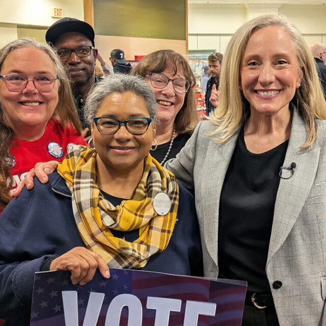 6 Moms Demand Action volunteers stand with Abigail Spanberger, who wears a black shirt, dark pants, and a light grey blazer. One volunteer holds a sign that says 