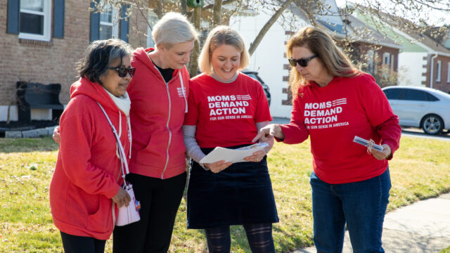 A group of four volunteers in Moms Demand Action shirts look at a piece of paper while door knocking