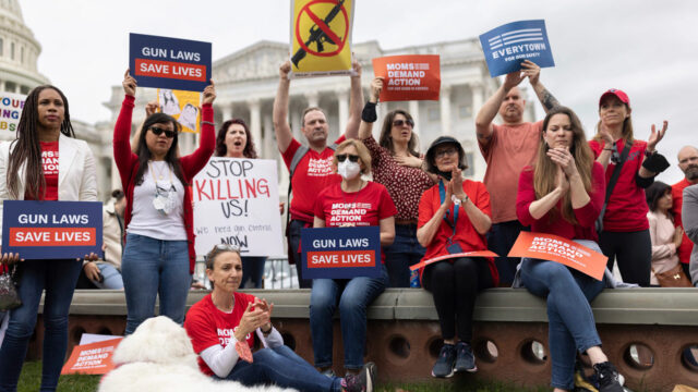 Moms Demand Action volunteers holding up a variety of signs: Gun Laws Save Lives, an assault rifle with a crossed out line through it, and Moms Demand Action and Everytown logo signs