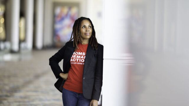Angela Ferrell-Zabala, the executive director of Moms Demand Action, poses for a portrait photo in the atrium of a building with lots of windows and natural light. She is wearing a black blazer, a red Moms Demand Action t-shirt, and blue jeans; she leans against a wall with her right hand on her hip while gazing slightly off the to the left.