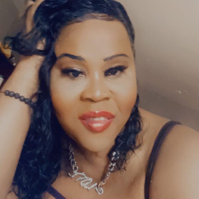 Monique Brooks, a Black trans woman, takes a selfie. Her right hand is scrunched into her hair, which is black, curly, and falls around her shoulders. She wear a beaded bracelet on her right wrist and a necklace with a chunky silver chain.