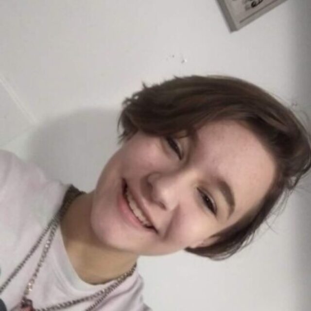 River Nevaeh Goddard, a white nonbinary person, takes a selfie at a 90-degree angle. River has short brown hair cut to just above their chin. They are wearing a light t-shirt and a necklace with a metal chain and a chunky gemstone.