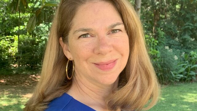 A woman with light skin and shoulder-length, light-brown hair is wearing gold hoop earrings and a cobalt blue, sleeveless blouse. She is softly smiling at the camera and is standing in what appears to be a backyard, with green trees and green grass visible in the background. She is pictured from the shoulders up; her torso is turned about 80 degrees away from the camera, and her head is directly facing the camera.