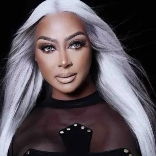 Vanity Williams, a Black trans woman, poses for a photo. Her hair is long, silver, and straight; it falls below her shoulders. She wears long eyelash extensions and a black top with a mesh neckline.