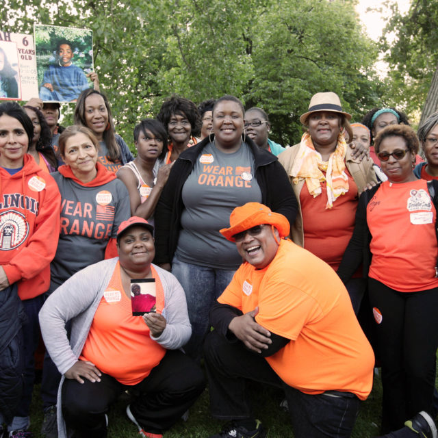 A group of people smile at a Wear Orange event. Some hold up signs for the loved ones they've lost to gun violence.