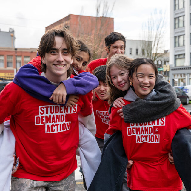 Six Students Demand Action volunteers stand in pairs, with one student per pair holding the other in a piggy-back position. They are all wearing red Students Demand Action t-shirts over top of sweatshirts or other warm clothes.