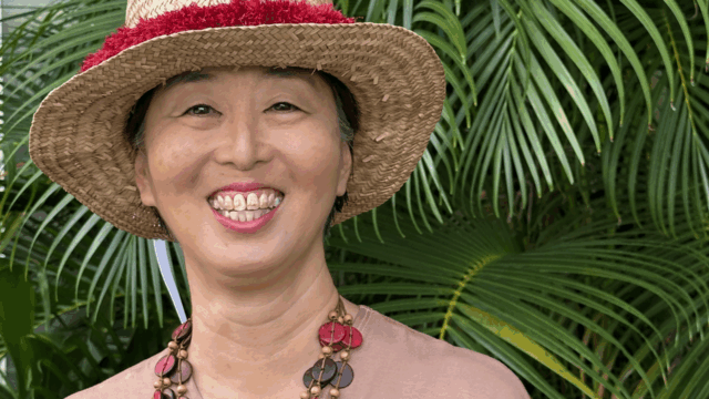 A photo of Elna Nagasako from the waist up, who is pictured in front of some ferns while wearing a dusky rose-colored pink wrap top, a chunky multi-strand necklace, and a straw hat.